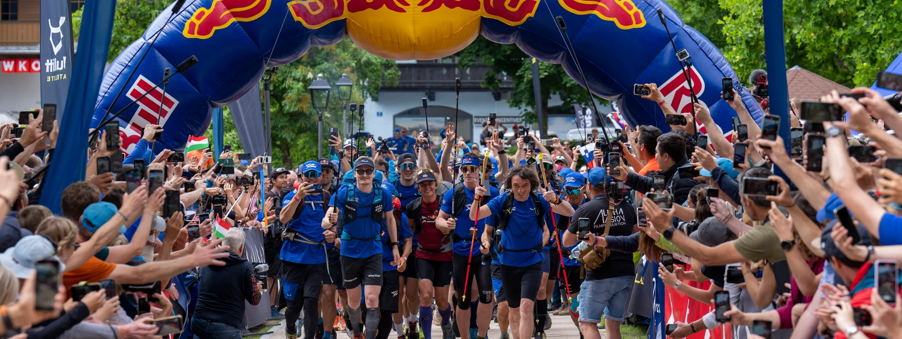 Partenza Red Bull X-Alps con atleti sotto arco gonfiabile e spettatori esultanti a Kitzbühel