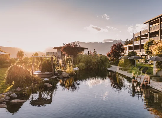 Naturteich bei Sonnenuntergang vor einem Wellnesshotel mit Liegestühlen, üppiger Bepflanzung und Bergkulisse