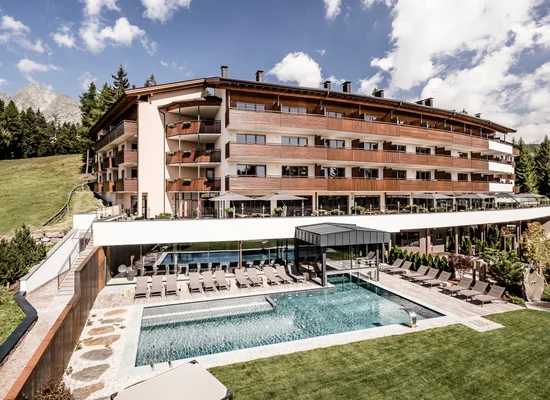 Berghotel mit Holzbalkonen, Außenpool und Liegebereich inmitten der Südtiroler Berglandschaft im Sommer